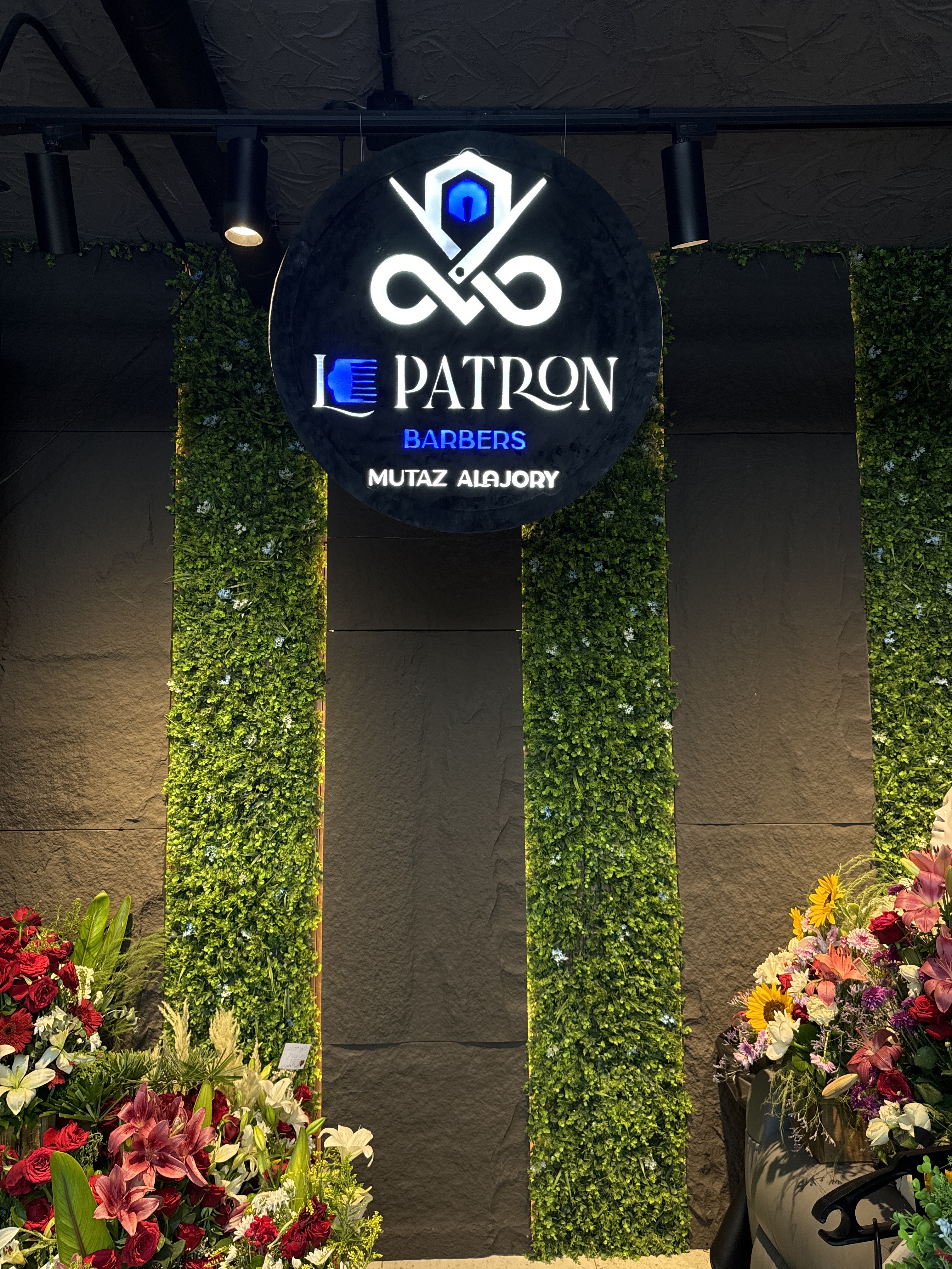 The illuminated Le Patron Barbers sign framed by a live plant wall and opening-day flowers