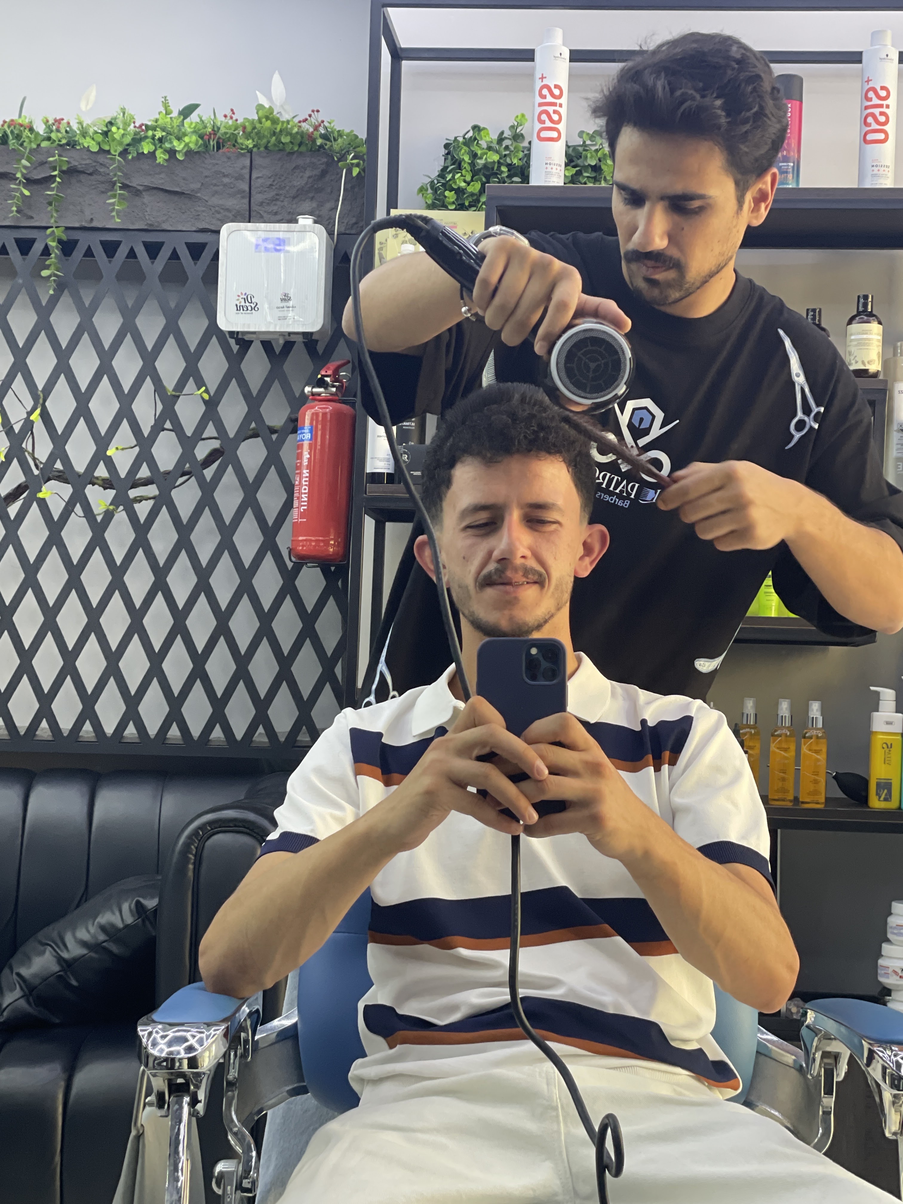 Barber finishing a client's style with a diffuser and comb beside shelves of styling products