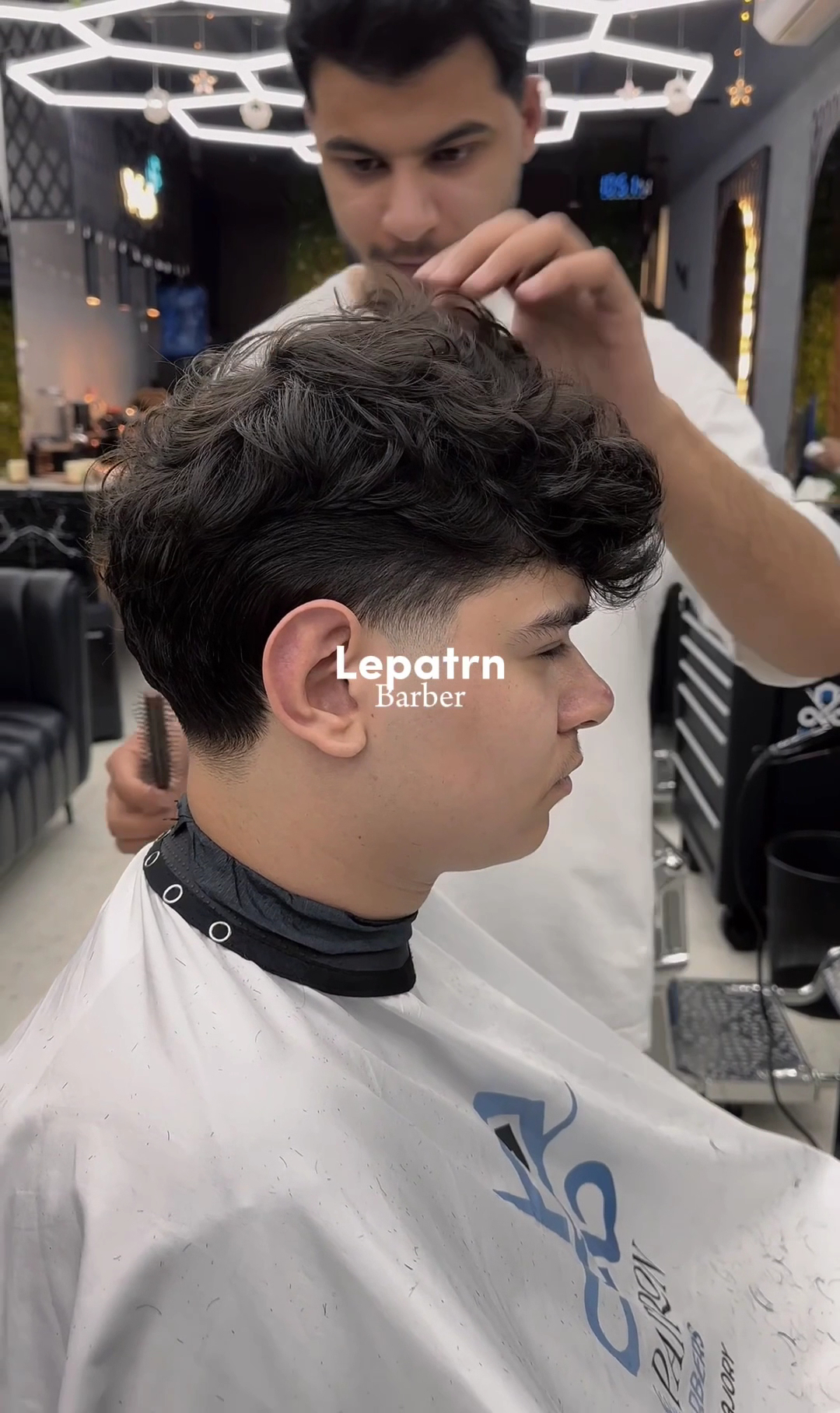 Barber shaping a client's fade and textured curly top under warm honeycomb lighting